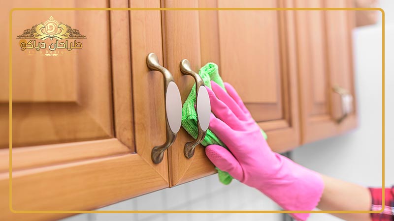 فردی در حال تمیز کردن کابینت چوبی با دستمال سبز و دستکش صورتی.